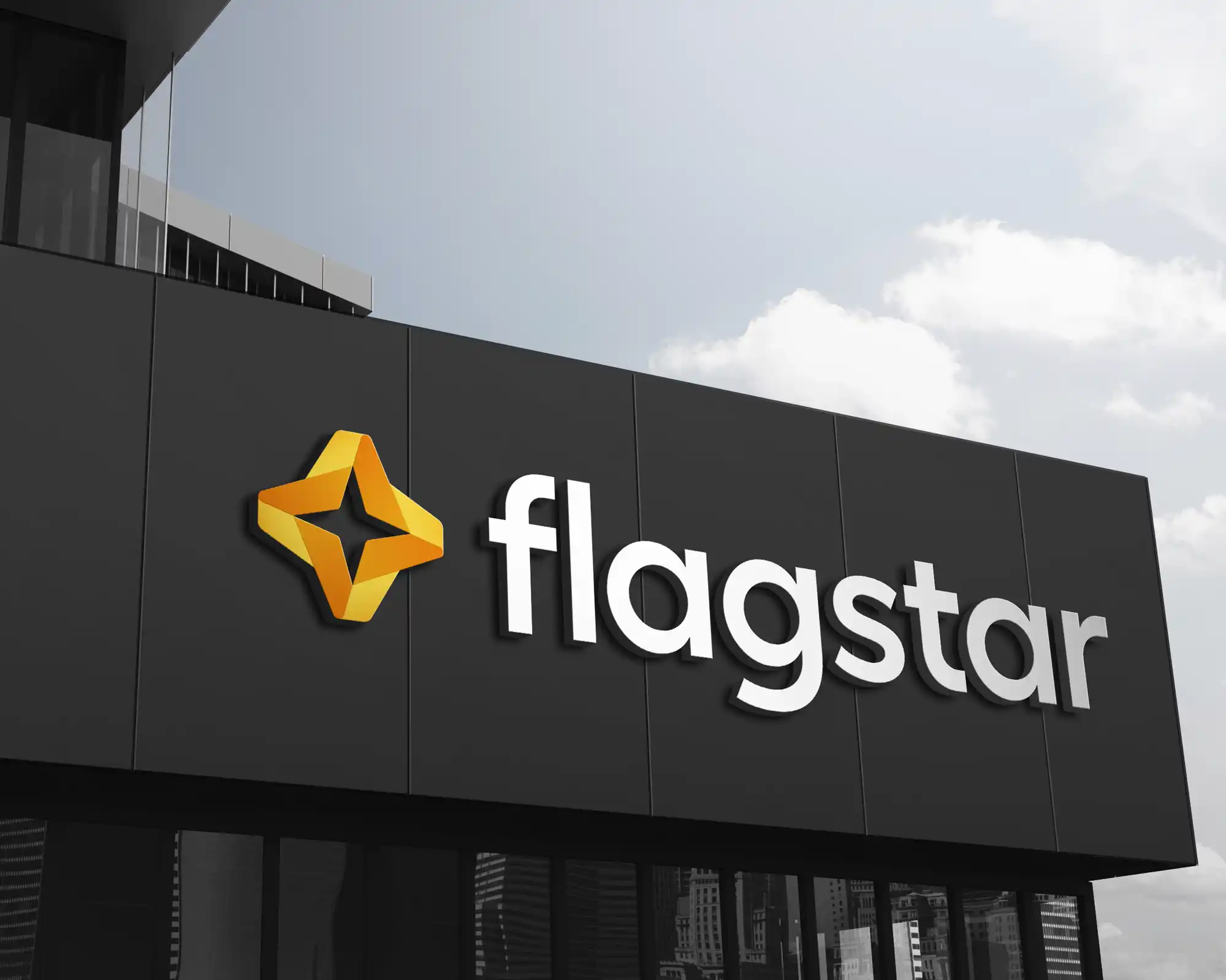 Modern building facade with dark gray vertical paneling featuring the Flagstar logo - an orange and yellow star with folded ribbon design - and white raised lettering spelling "flagstar" against a blue sky with scattered clouds.