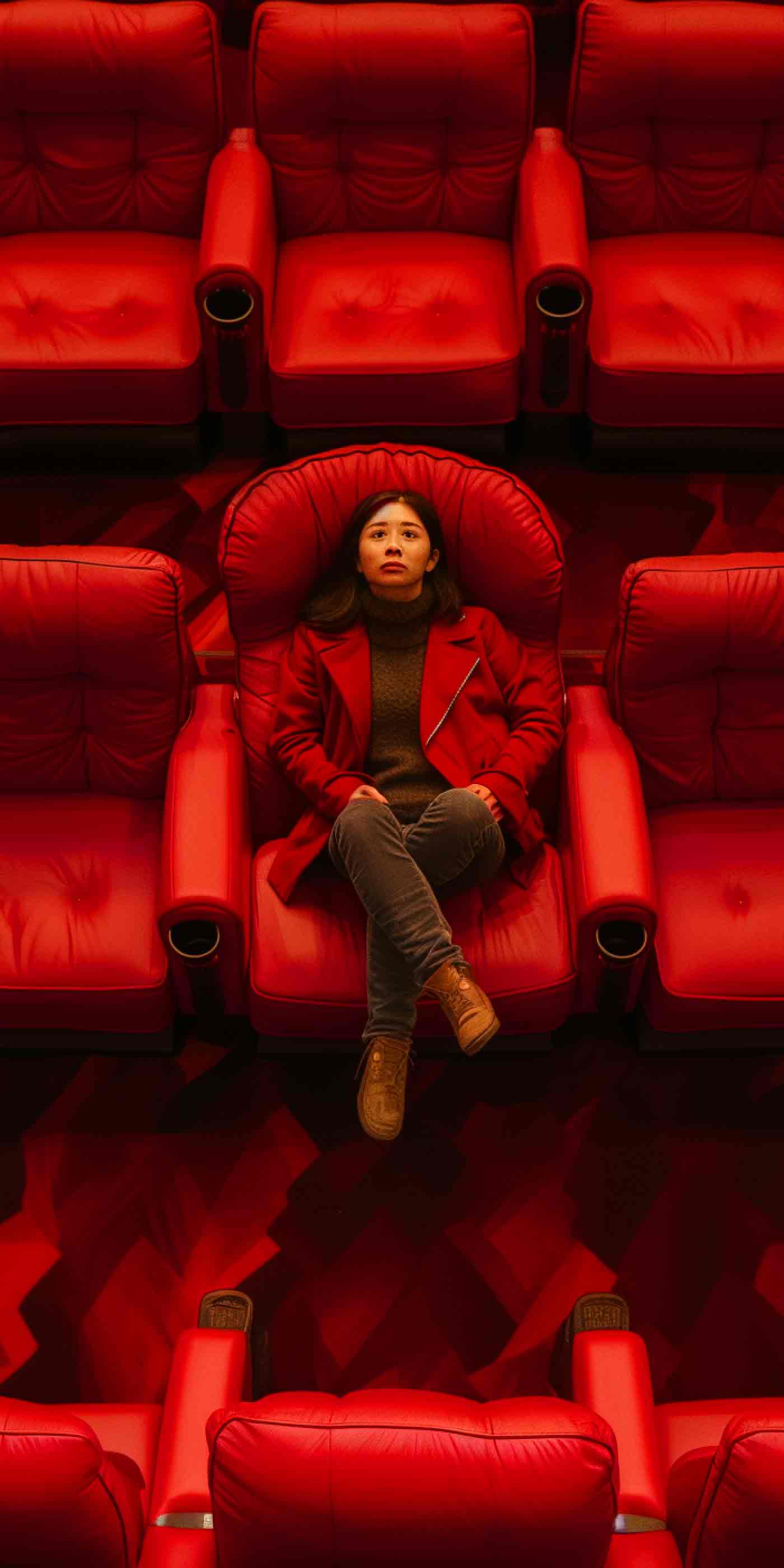 Young woman in a red coat sitting alone in an empty movie theater, looking upward pondering the desire for creative and movement marketing insights.