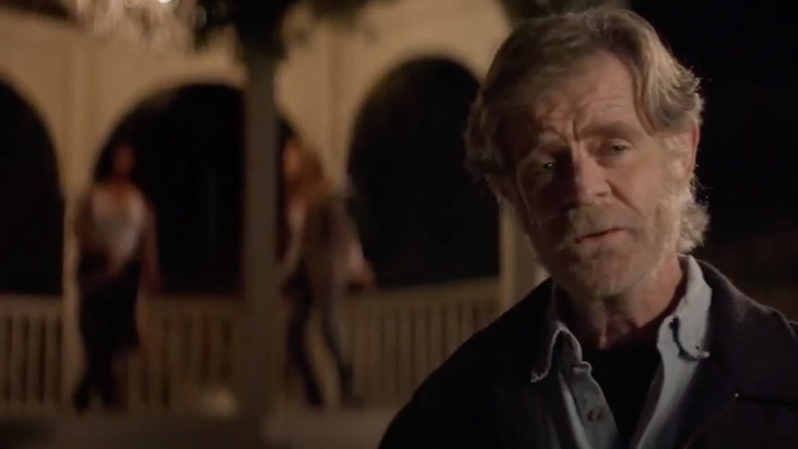 Close-up on actor William H. Macy looking off-camera while two people are blurred in the background. 