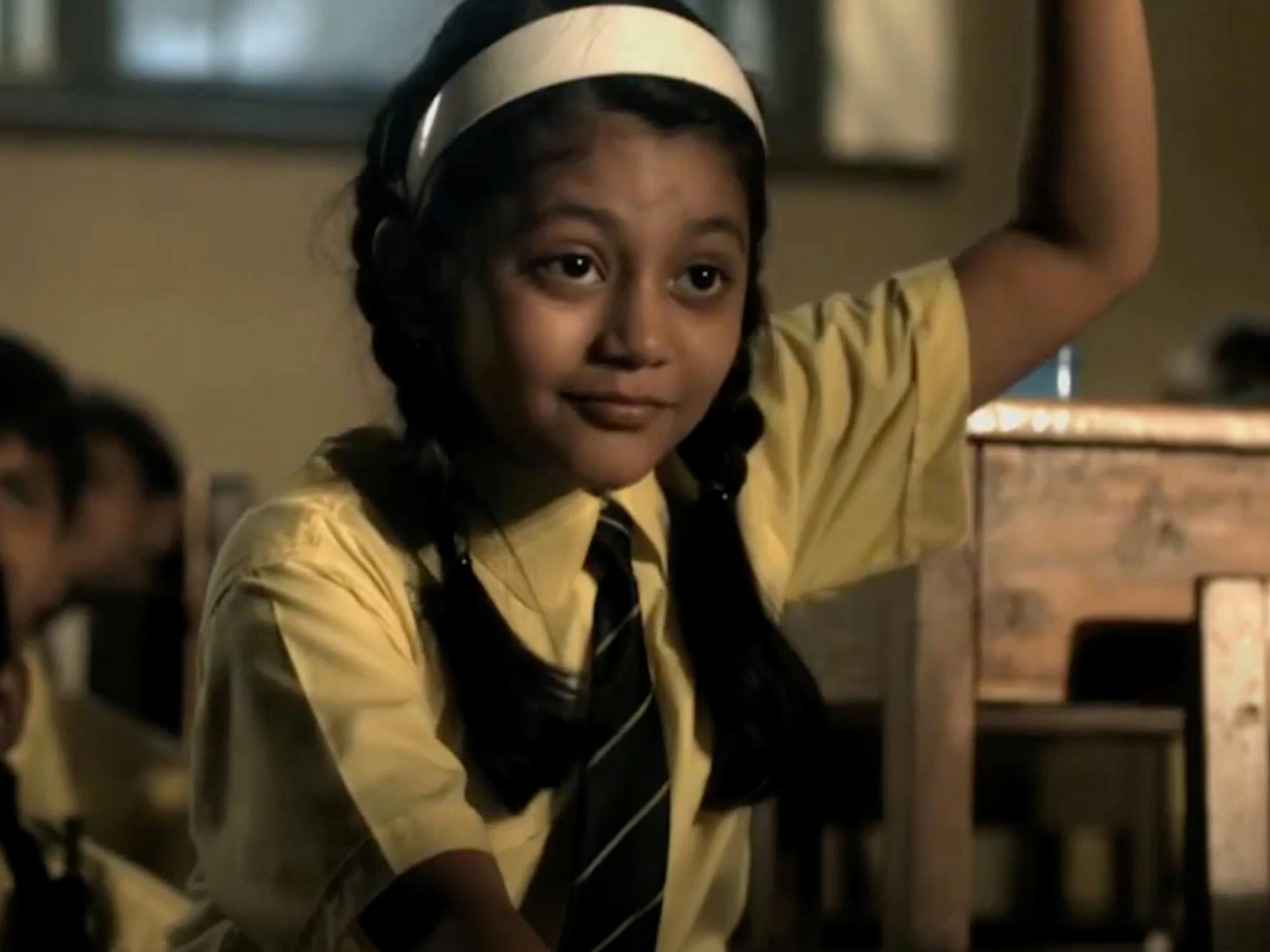 Close-up of a young schoolgirl in uniform and pigtails raising her hand inside a classroom.